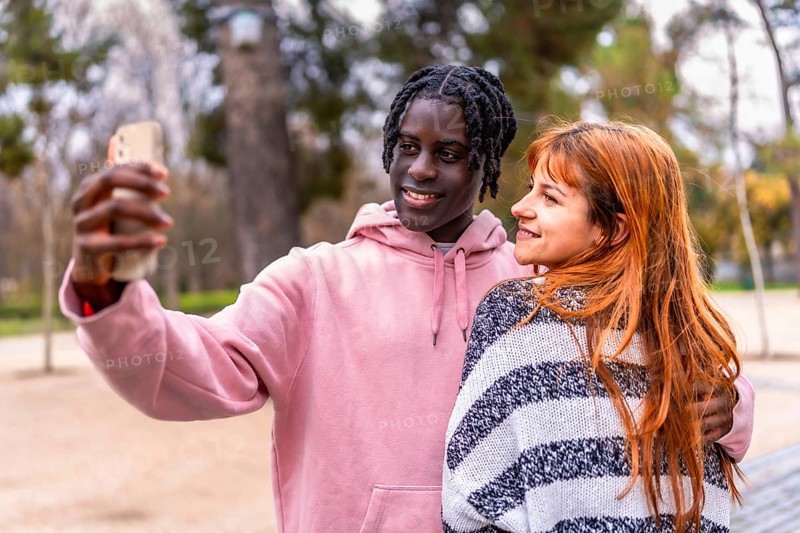 Young multiracial couple smiling together while capturing a joyful selfie with a smartphone in a vibrant park setting