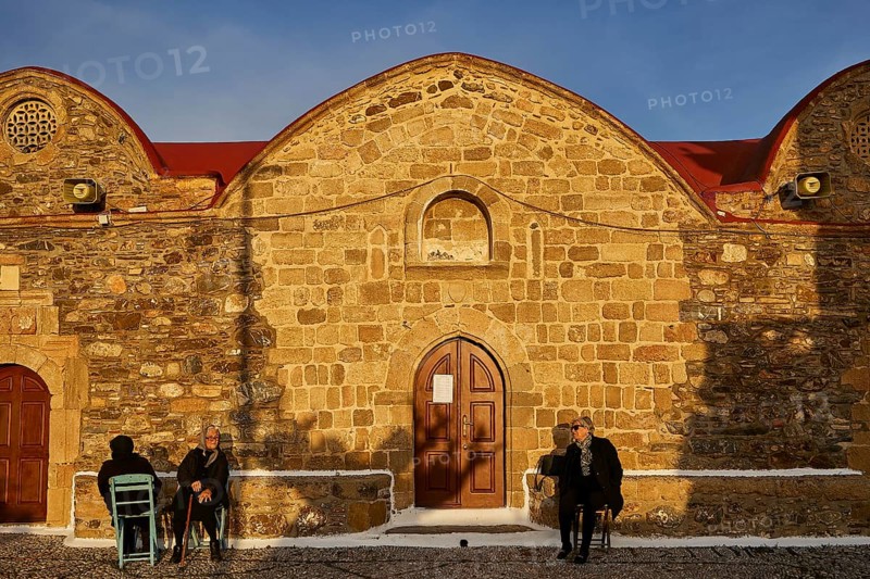 People sitting in front of a stone church in warm light and quiet surroundings, Easter 2025, Church of the Dormition of the All-Holy God Bear, Kimisis tis Theotokou, Asklipio, Rhodes, Dodecanese, Greek Islands, Greece, Europe