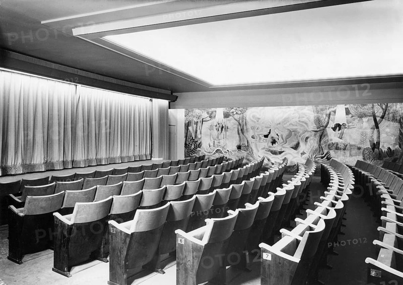 Emtpy chairs. A picture from a movie theatre with empty seats. The rows of chairs are numbered so the people coming with tickets and seat details can find their place in the theatre. The curtain covers the film screen. The wall is decorated. Sweden 1950s