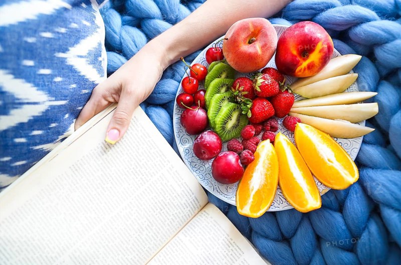 Plate with fresh fruit salad on a blue plaid and book. Woman reading the book and eating fresh fruits. Close up. Top veiw. Cozy and healthy concept