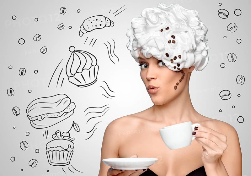 Young beautiful woman with whipped cream on her hair, with a curl of coffee beans, drinking coffee and holding a coffee cup, against a sketchy background of bakery sweets