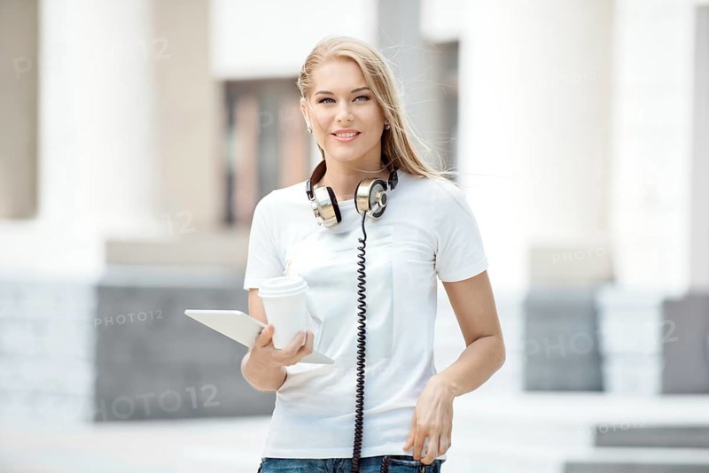 Happy young woman with vintage headphones around her neck, holding a cup of coffee to go and surfing the internet on a tablet PC, against an urban background