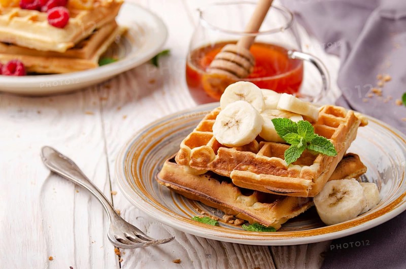 Belgian waffles served with banana and mint leaf on white wooden kitchen table with syrup aside