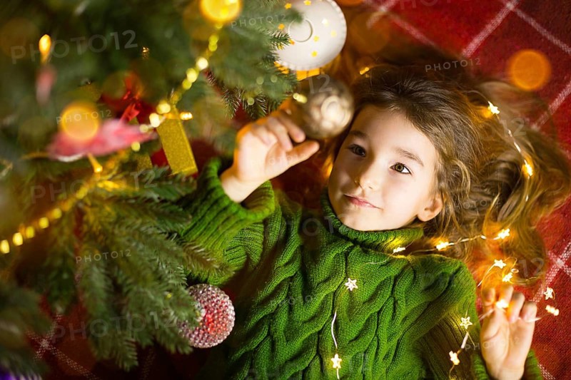 A girl with long hair and garlands lies on a red plaid under a Christmas tree with toys in a warm knitted sweater. Christmas, New Year, children's emotions, joy, expectation of a miracle and gifts