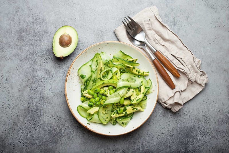 Healthy vegan green avocado salad bowl with sliced cucumbers, edamame beans, olive oil and herbs on ceramic plate top view on grey stone rustic table background, food photography, food photography