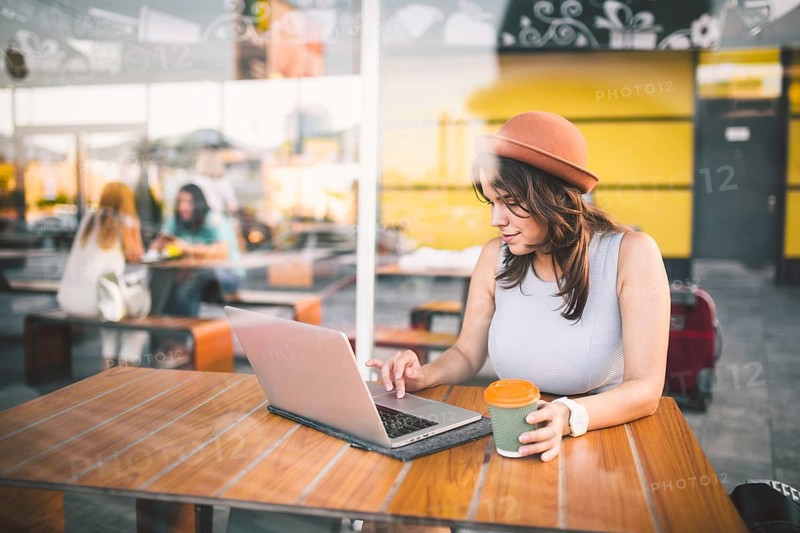 Beautiful young caucasian woman sitting on a terrace in a cafe in summer at a wooden table in a hat and a plate uses technology, working behind a laptop booking hotels trip in the summer