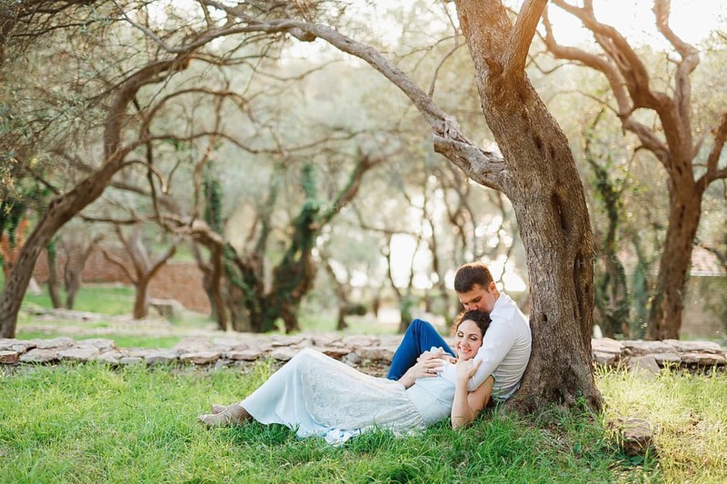 Kotor, Montenegro - 21.06.17: Seated man hugs a woman lying next to him on green grass and kisses her on the crown near a tree in an olive grove