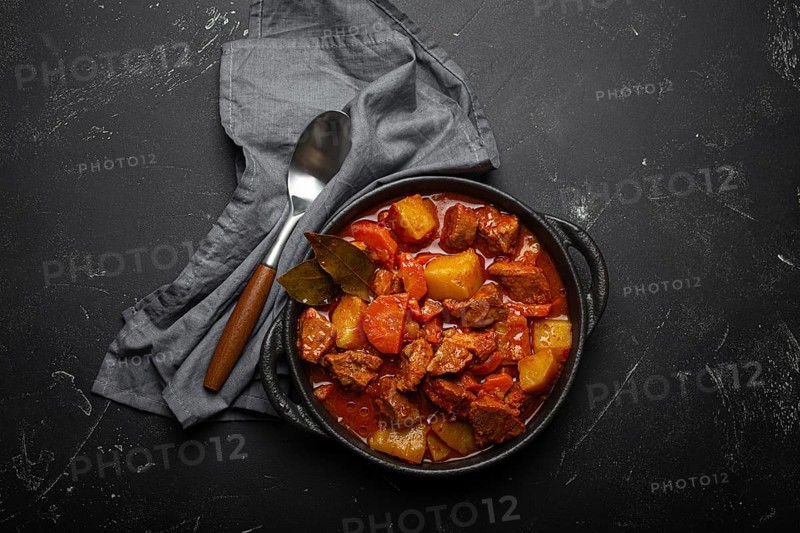Beef meat stew with potatoes, carrot and delicious gravy in black casserole pot with bay leaves with spoon on black dark rustic concrete background from above, food photography
