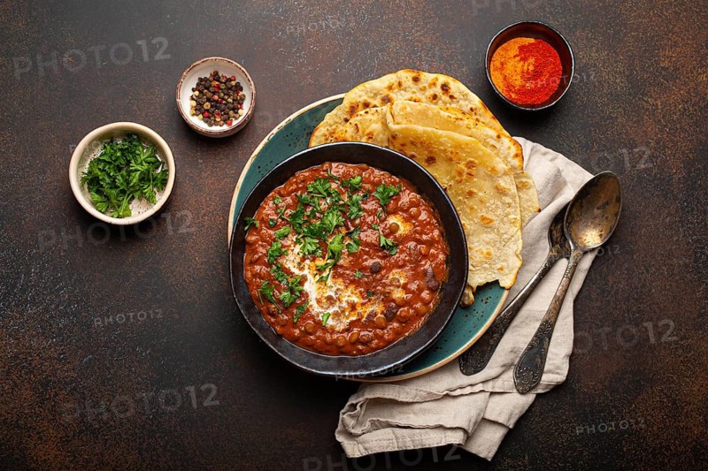 Traditional Indian Punjabi dish Dal makhani with lentils and beans in black bowl served with naan flat bread, fresh cilantro and two spoons on brown concrete rustic table top view, food photography, food photography