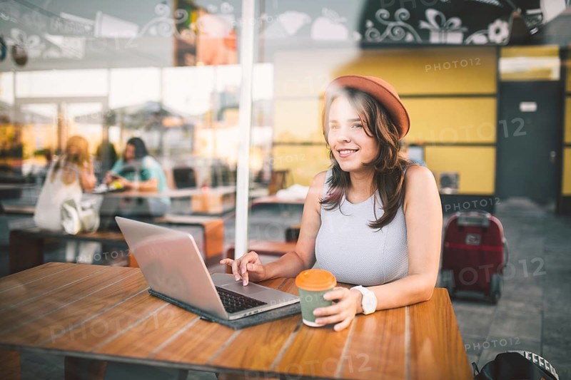 Beautiful young caucasian woman sitting on a terrace in a cafe in summer at a wooden table in a hat and a plate uses technology, working behind a laptop booking hotels trip in the summer