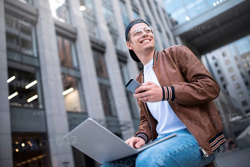 Happy handsome student looking at computer screen while typing something on laptop keyboard during work break outdoors, teenager looking at the notebook sitting at street