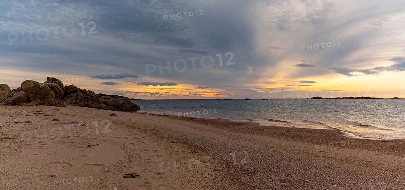 A panorama view of a golden sand beach at sunset with calm ocean water and red granite rocks