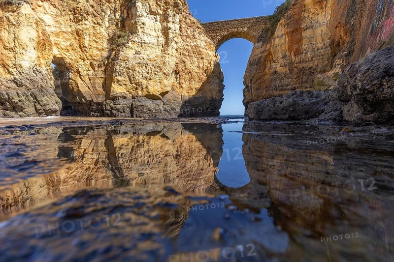 Yellow rocks and cliffs on a sandy beach. A bay by the sea, over which the old stone bridge of the Pinhão fortress connects two cliffs. Praia da Caldeira, Lagos. Algarve, Portugal, Europe