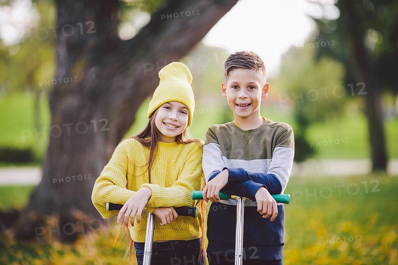 Two children caucasian boy and girl twins posing with scooters in autumn park. Happy kids riding kick scooters in public park. Kids sport. Outdoor activities brother sister teens. Happy childhood