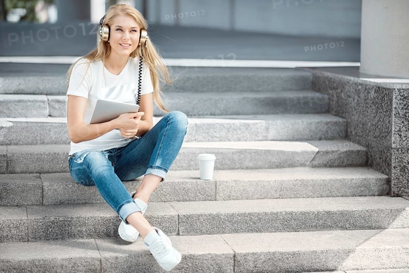 Happy young woman with vintage headphones and a cup of coffee to go, surfing the internet on a tablet PC, listening to music and sitting on a staircase in front of an urban background