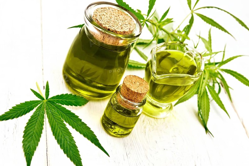 Hemp oil in two glass jars and sauceboat, leaves and stalks of cannabis on the background of wooden boards