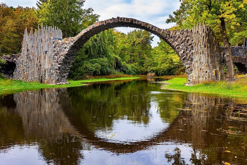 The Devil's Bridge on the Rakots River. The basalt columns of the bridge form a perfect circle with their reflection in the water. Germany. Picturesque narrow bridge in the park Kromlau