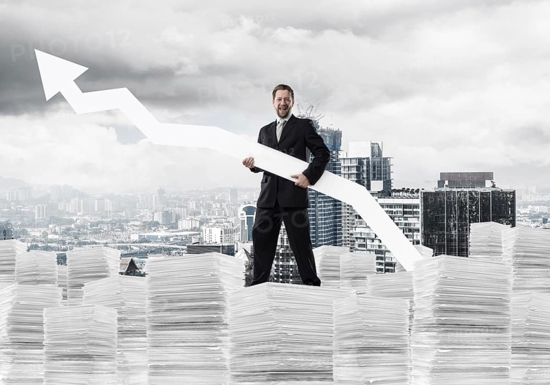 Young and ambitious businessman in suit holding big white arrow in hand and pointing away while standing on paper column