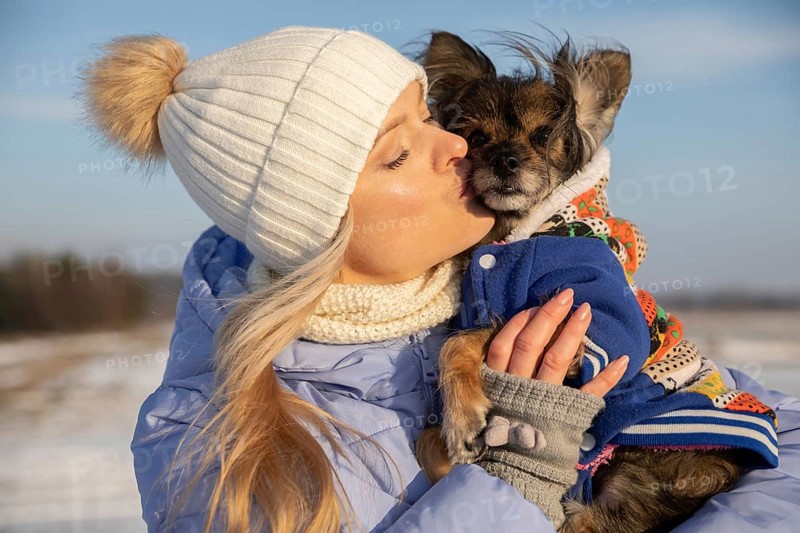 Close-up of a blonde woman hugging her dog. The woman and dog are outside on a winter day. Both are dressed in warm coats