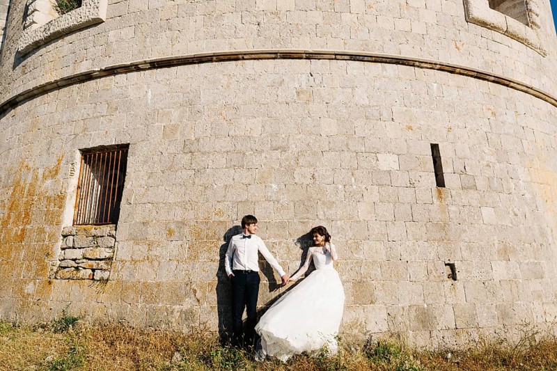 Bride and groom are holding hands and standing tjgether near the wall of the Arza fortress.