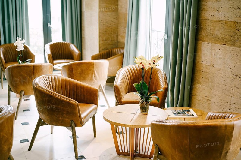 A large hall with exits to the balcony with round tables and flowers and brown armchairs made of genuine leather.