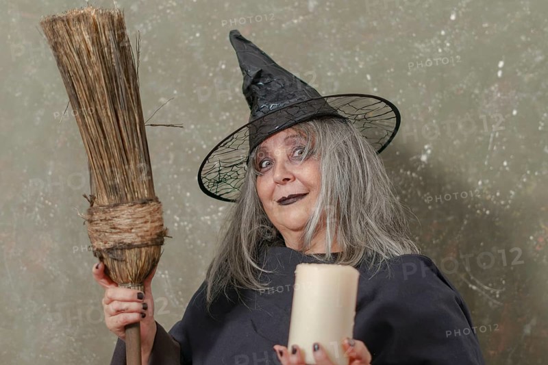 Old witch with white hair and a black hat holding a broom in her hand and a candle in the other while smiling