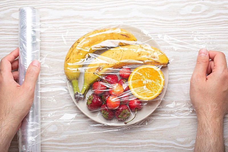 Male hands wrapping assorted different fruit and berries on white plate in plastic clear film top view on white wooden rustic table background, food photography, food photography