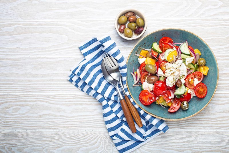 Greek fresh healthy colorful salad with feta cheese, vegetables, olives in blue bowl on rustic white wooden background top view, Mediterranean diet, traditional cuisine of Greece. Space for text, food photography
