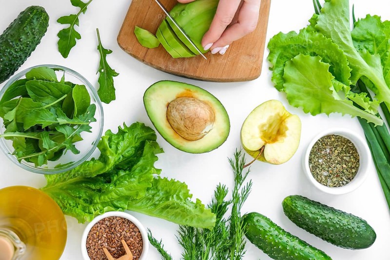 Woman cooking salad of fresh green vegetables and herbs. Cooking healthy diet or vegetarian food. Female hands cut avocado on cutting board surrounded by green vegetables. Step by step recipe. Healthy eating. Ingredients for salad. Pattern flat lay with healthy vegetarian meal ingredients. Raw food concept. A variety of organic fruits and vegetables with avocado. Vegan menu. Fresh spinach and green onion leaves