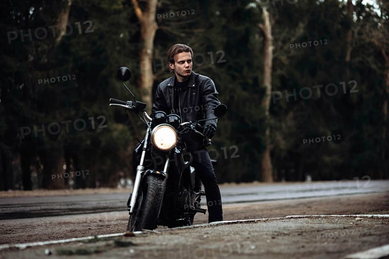 A person in a leather jacket sits on a motorcycle by a forest road, creating a moody atmosphere