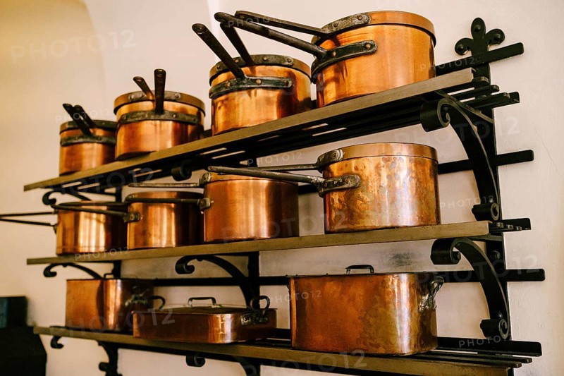 Pots, ladles, tray and saucepan made of copper, in various sizes, on massive metal shelves, fixed to a white wall.