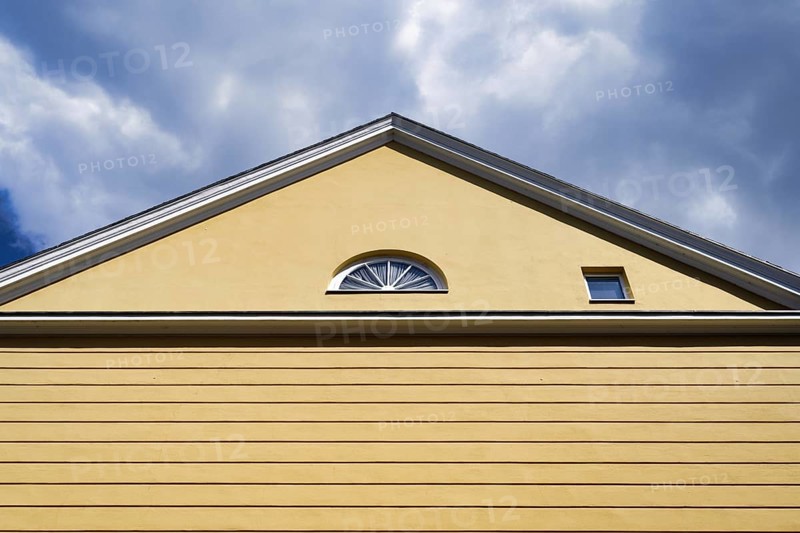 A yellow house with a pitched roof and one semi-circular and one square window in front of a cloudy sky semi-circular window