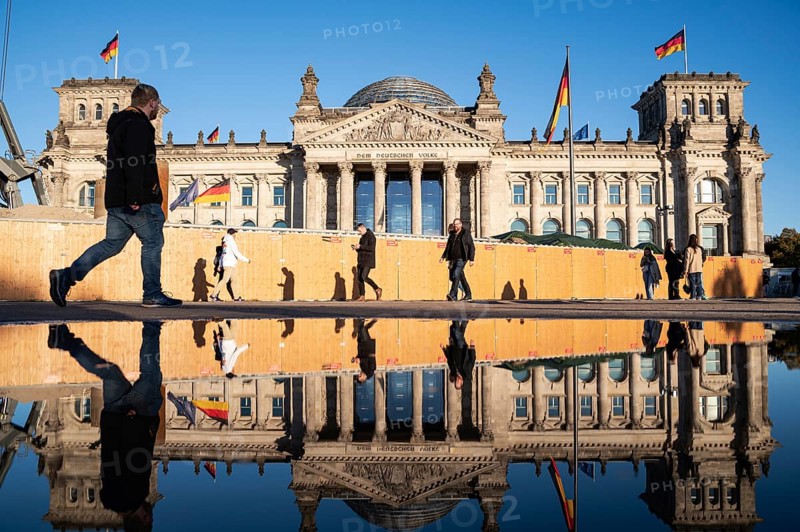 21.10.2023, Berlin, Germany, Europe, People walk past the Reichstag in Berlin's Mitte district on a sunny autumn day, its west façade reflected in a large puddle in front of it. On the façade of the Reichstag building above the portal is the lettering, Europe