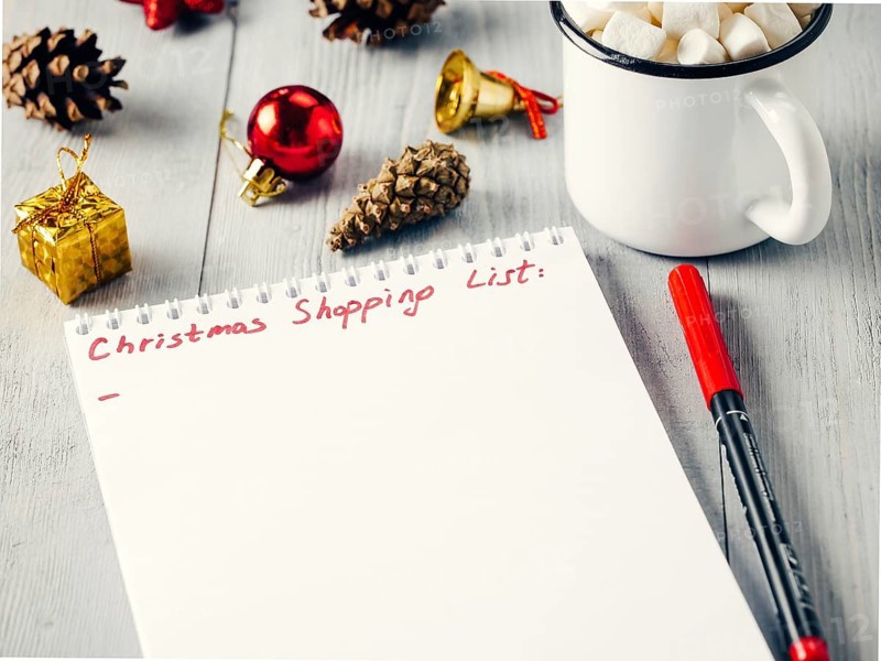 Christmas gifts shopping planning. Make shopping or to-do list for Christmas. Notebook, mug hot chocolate with marshmallows, New Year's decoration and pine cone on gray wooden background