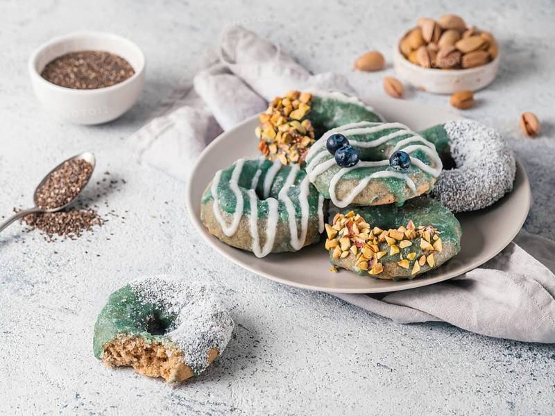 Vegan doughnuts with chia seeds topped with healthy spirulina glaze with pistachio, desiccated coconut and blueberry. Blue green spirulina donuts on gray background. Copy space