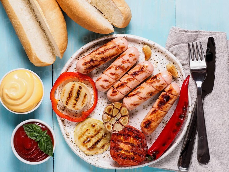 Top view of chicken homemade sausages, buns bread, sauces ketchup and mustard on blue wooden background. Grilled sausages and grilled vegetables in craft trendy plate. Homemade hotdogs