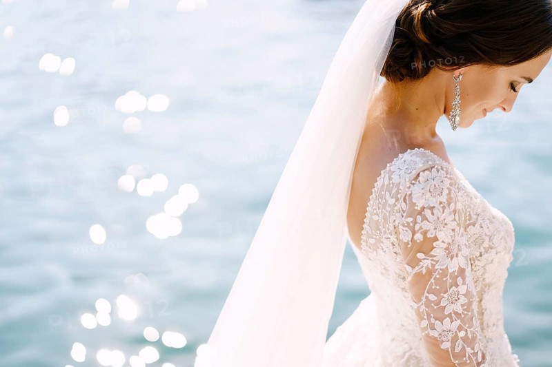 Close-up of the neck and neck of the bride, face profile. Beautiful big earring in ears, shoulder with white lace dress on background of glare of water. Fine-art wedding photo in Montenegro, Perast