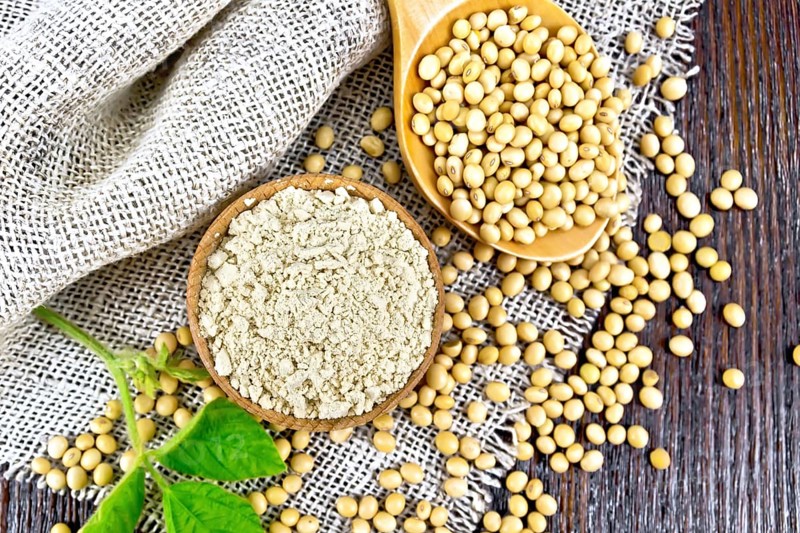 Soy flour in the bowl, soybeans in a spoon and on sackcloth, green soya leaf on a wooden board background from above