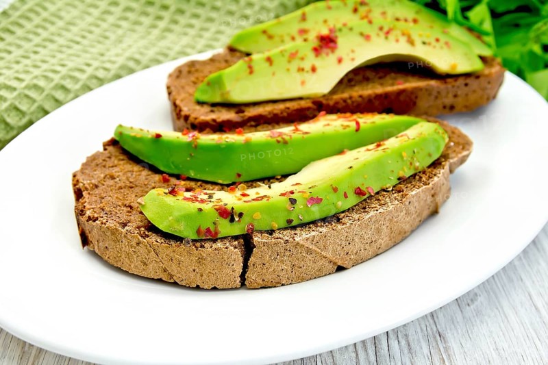 Two slices of rye bread with slices of avocado and pepper on a plate, green cloth, parsley on the background light wooden boards