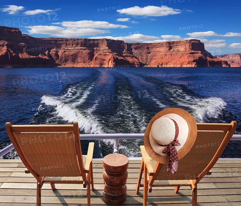 Waves from the boat cut through the Lake Powell. At the stern of the vessel are two deck chairs. On the back of one hanging elegant ladies straw hat