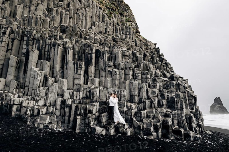 A wedding couple stands on a wall of stone pillars. The bride and groom are hugging on basalt Kekurs, on the black sandy beach of Vik. Destination Iceland wedding