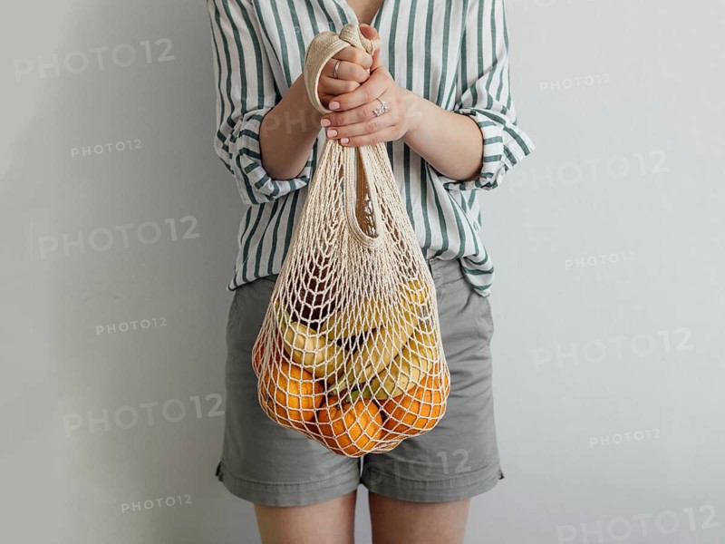 Mesh bag with fruits in female hands. Stylish young woman hold mesh shopping bag on light gray wall. Modern reusable shopping concept