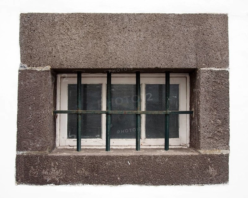 A small white wooden window with glass panes covered by steel bars in a large grey stone frame