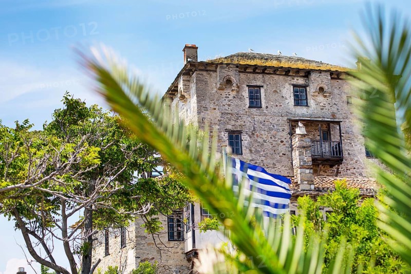 Ancient Ouranoupolis Tower on Athos peninsula in Halkidiki, flag of Greece. View through the palm leaves