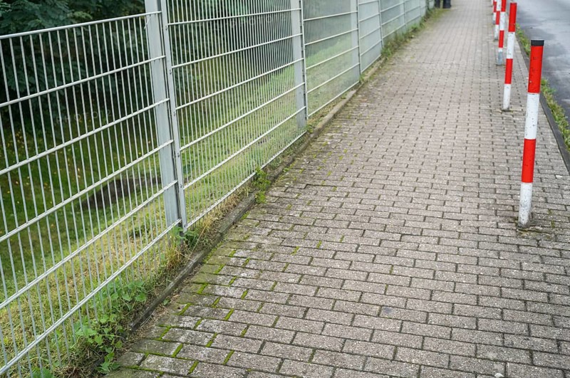 Sidewalk, metal fence on the left, red and white poles on the right