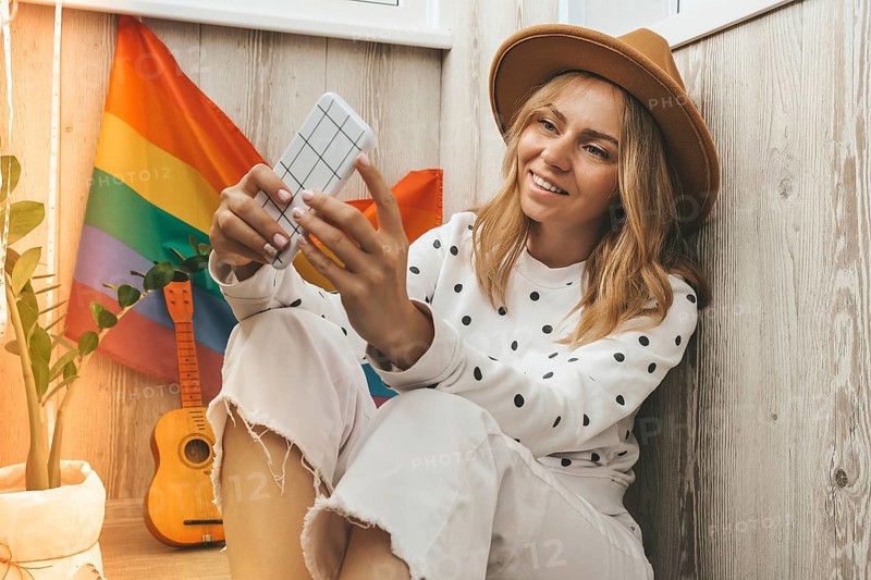 Young millennial hippie woman sitting on balcony using mobile phone. LGBTQ rainbow flag on background. Phone texting, video call, taking selfie. Blogger. Work from home. Modern office. Blonde hair. Stay home