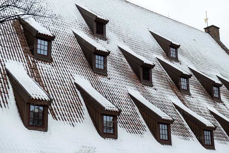 Snow falling on the roofs of the city. Snow-covered brown metal tiles roof of European house with windows. Rooftop covered in fresh snow. Winter scene