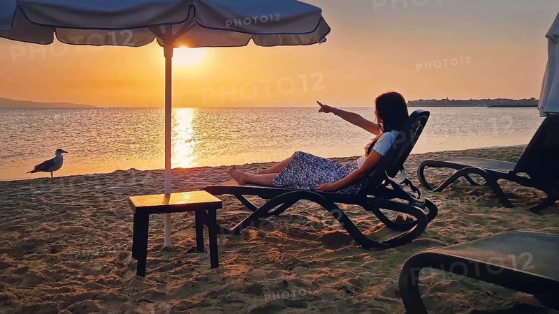 Young woman sitting on a sunbed at the beach pointing forefinger to the sunrise over the horizon. Beautiful female taking a sunbath at the sea. Summer vacation seaside, holiday relish concept