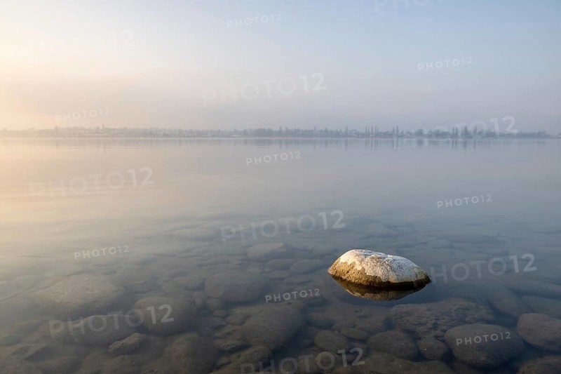 A large stone rests in the calm waters of a misty lake at dawn, harbour, Allensbach, Baden-Württemberg, Germany, Europe