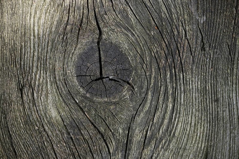 Detailed view of a piece of aged wood with a gray-brown coloration that emphasizes its natural textures and patterns. Deep cracks and knots cover the surface